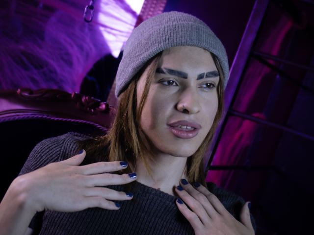 AaronFinn has light skin and brown hair, wearing a gray beanie and a dark gray rib-knit sweater. His eyebrows are thick and arched. He has dark blue nail polish on his fingers, which are placed near his neck. His lips are slightly parted, showing a subtle, relaxed expression. The image uses purple and pink lighting, creating a contrast with his gray clothing. The background is blurry, featuring metallic and textured elements. The photograph focuses on AaronFinn's face and upper body.