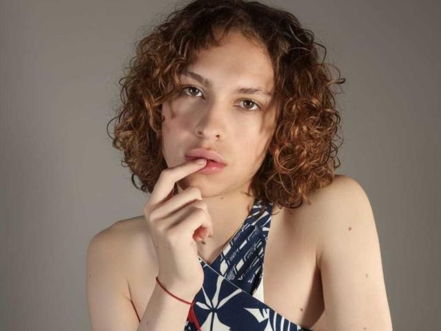 AaronFinn, com cabelo castanho encaracolado, pele clara e lábios rosados, mordendo o dedo, vestindo uma blusa de alça azul e branca, fundo cinza, pulseira vermelha no pulso..