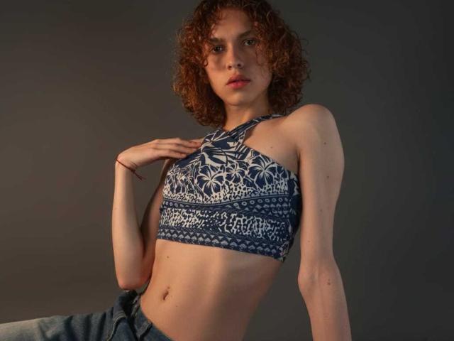 AaronFinn with curly brown hair, wearing a blue patterned sports bra, showing a slim flat stomach and small tits, with blue jeans, hand on chest, looking at camera.