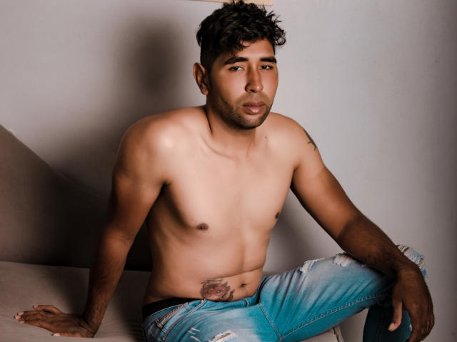 DantteRay, shirtless with a toned, brown-skinned body, small nipples, and a tattoo on his right hip, wears torn blue jeans, sitting on a couch, looking at the camera.