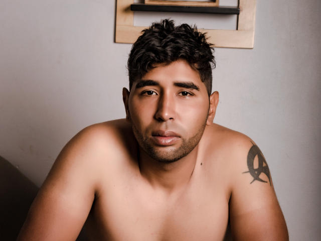 Shirtless DantteRay with curly black hair, light brown skin, and a black tattoo on his right shoulder, staring at the camera with a neutral expression.