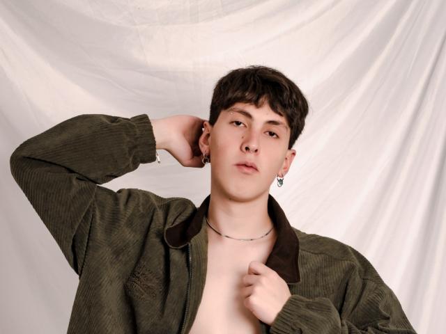 MilanStone, a young dude with short brown hair, wearing an unzipped green cord jacket, hand in hair, earring, and necklace, looking slightly bored.