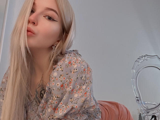 StellaMoor with long blonde hair, pouty lips, wearing a floral shirt, and pink pants, leaning forward, showing her small tits, in a slightly angled selfie.