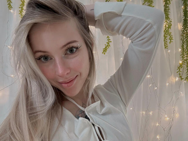 StellaMoor with long blonde hair, blue eyes, and light makeup, wearing a white ribbed shirt, hand in hair, smiling, soft fairy lights in background.