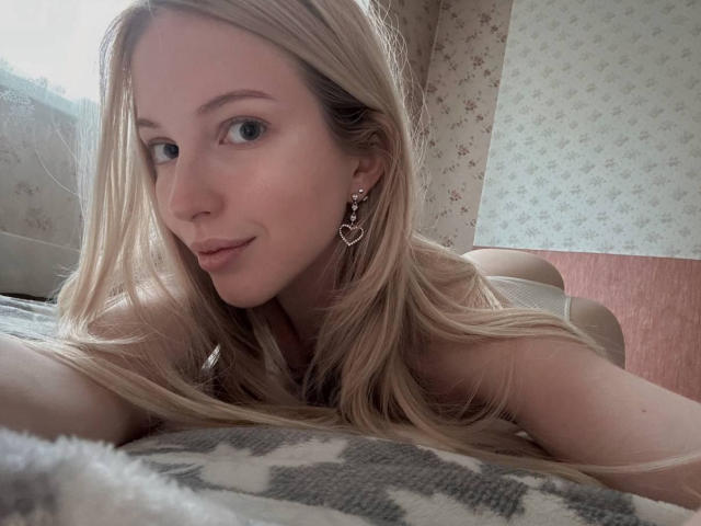 StellaMoor with long blonde hair, blue eyes, and heart-shaped earrings, lying on a patterned blanket, smiling softly, showing her bare shoulders and upper chest.