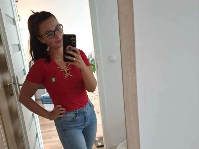 CuteLexy-hot with glasses, red top, gold necklace, and blue jeans, taking a mirror selfie, pouting, showing her ass, standing in a doorway, holding a black phone.