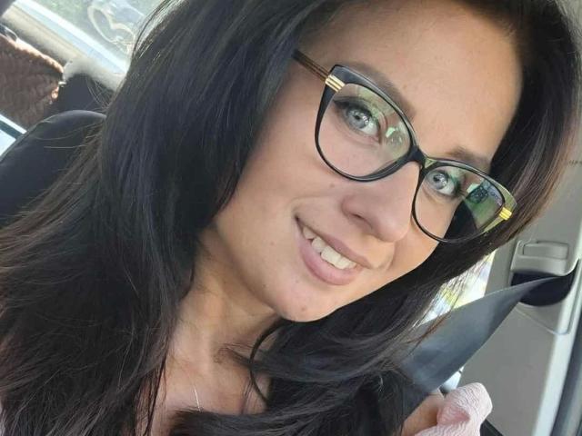 CuteLexy-hot with dark hair, glasses, and a seatbelt, smiling in a car, showing her teeth, holding the belt with a pink hand. Her tits are partially visible.