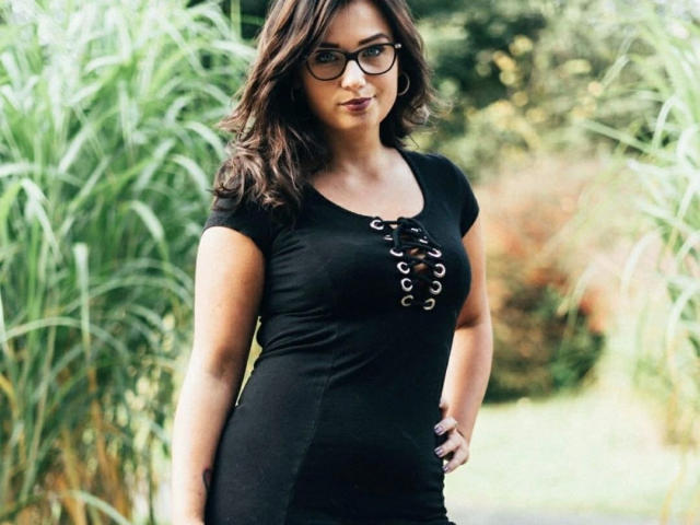CuteLexy-hot with glasses, wavy brown hair, black lace-up top, showing her tits, standing in front of green reeds, hand on hip, smiling, outdoors, daytime.