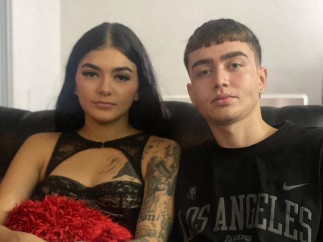 SexyFlaquitaIvi with black lace dress, showing her tits, holding red pillow, tattooed arm, and brown-haired dude in black "LA" Nike shirt, both sitting on black couch.