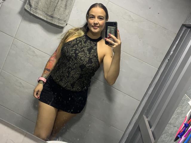 SaritaCaramelo stands in a bathroom, taking a mirror selfie. She has long blonde hair and wears a black lace sleeveless top with a high neckline and a black mini skirt. Her right arm, covered in a red and black tattoo, rests at her side. She holds a black phone in her right hand. Her pink smartwatch is on her left wrist. She has light skin and a slight smile. The mirror shows some water spots.