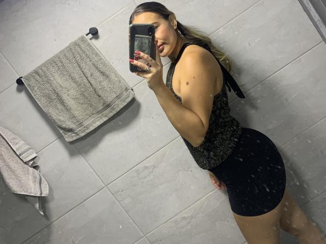 SaritaCaramelo with wet, blonde hair in a black, sleeveless, sparkly top and black shorts, taking a mirror selfie showing her curvy ass and small tits.