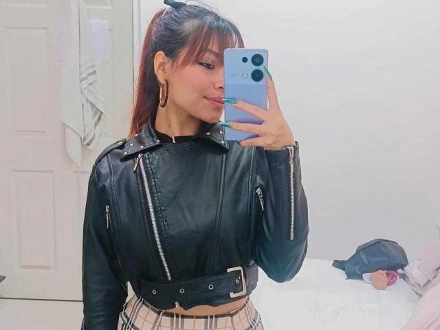 VannesaSaenz with red ponytail, wearing black leather jacket with zippers, striped skirt, and gold earrings, taking selfie in mirror, showing off green nails and subtle smile.