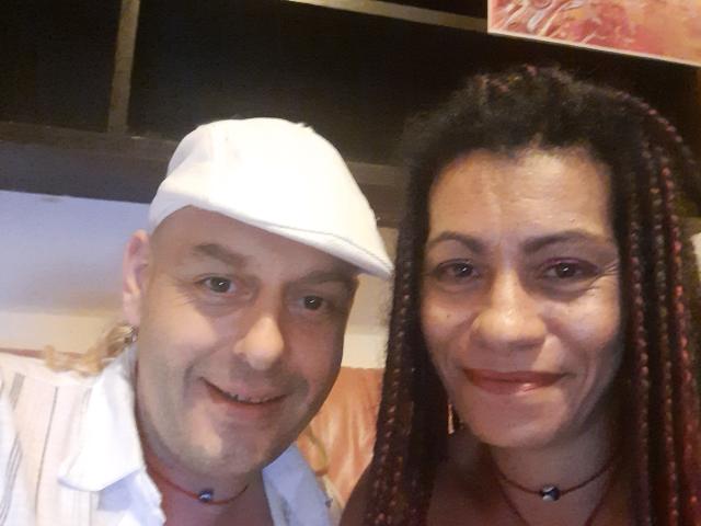 EtnikSatyr com a cabeça raspada e um boné branco, sorrindo, vestindo uma camisa listrada e um colar. EtnikSatyr tem cabelo trançado escuro, batom vermelho e também um colar..