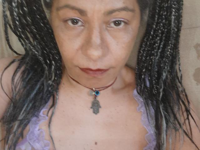 EtnikSatyr, avec des cheveux noirs et blancs tressés, une peau brun clair, portant un haut en dentelle violette et un collier rouge avec un pendentif en pierre bleue et une plume. Les seins d'EtnikSatyr sont visibles..
