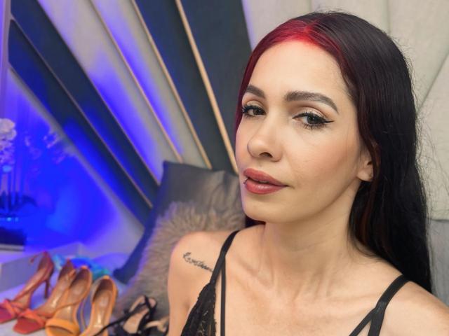 KathySteven, con capelli scuri, riflessi rossi, eyeliner nero e rossetto rosa, indossa una camicetta di pizzo nero, mostrando un tatuaggio sulla spalla destra, e un paio di scarpe con il tacco arancioni..