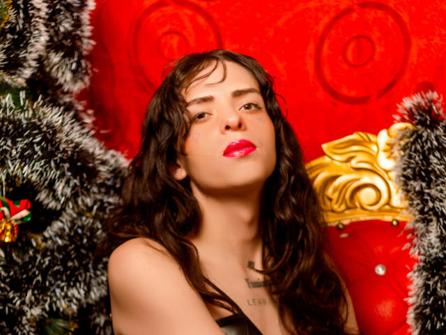 SashaSexiest with wavy brown hair, red lipstick, and a black top, standing between fluffy black and white garlands, looking up with a slight smile. Boobs visible, chest tattoo.