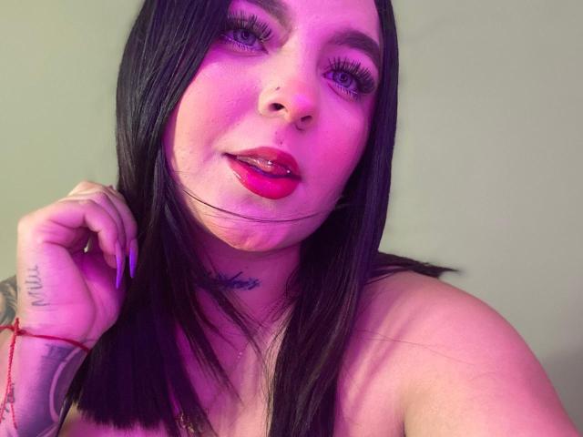 MelodyBlossom, con i capelli neri, il rossetto viola e un piercing al naso, ha un tatuaggio sul collo e un braccialetto rosso, e guarda la fotocamera con un leggero sorriso..