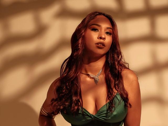 ScarlettFlever with red, wavy hair, green dress with deep cleavage, showing her tits, wearing a silver necklace and arm bracelet, in warm, shadowed light.
