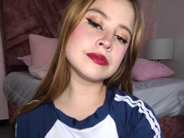 Melani Becker mit rotem Lippenstift, blonden Haaren und Eyeliner, die ein blau-weißes Sportshirt trägt, blickt leicht zur Seite und hat einen leicht schmollenden Gesichtsausdruck..