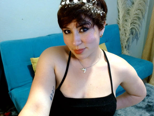 SaraKleine mit kurzen, braunen Haaren, schwarzem Top mit Halter, Schmetterlingskette, kleinem Tattoo am linken Arm, blauem Sofa und Blumenhaarreif, lächelt in die Kamera und zeigt ihre Brüste..