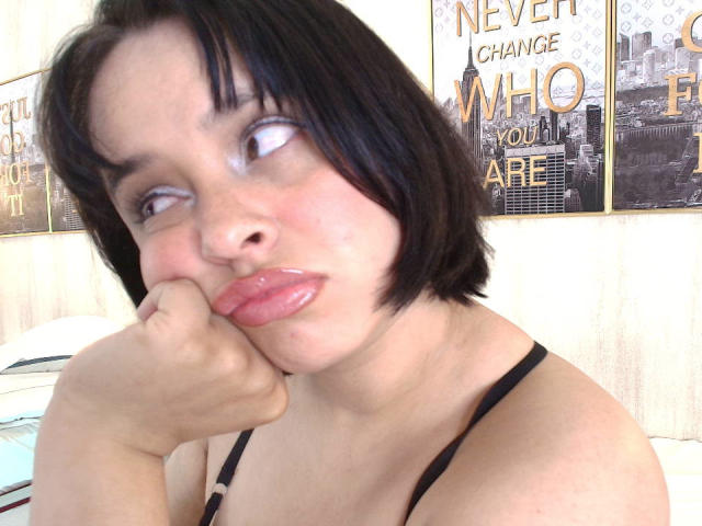 Blueeskyy with short black hair, glossy pink lips, and a pouty expression, resting her chin on her hand, wearing a black strap, looking to the side.