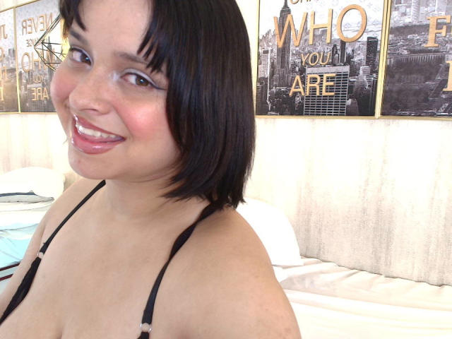 Blueeskyy, com cabelo preto curto e batom rosa, sorri para a câmera, mostrando seus pequenos seios em um top preto com tiras, sentada em uma cama com travesseiros brancos..