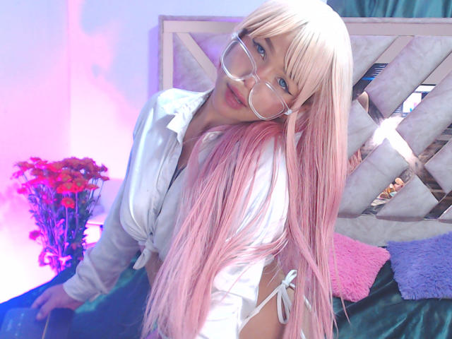 LuluDeVine with long pink hair, wearing glasses, white shirt, and side-tie bikini bottom, leaning forward, showing her ass, in a brightly lit room with pink and purple lights.