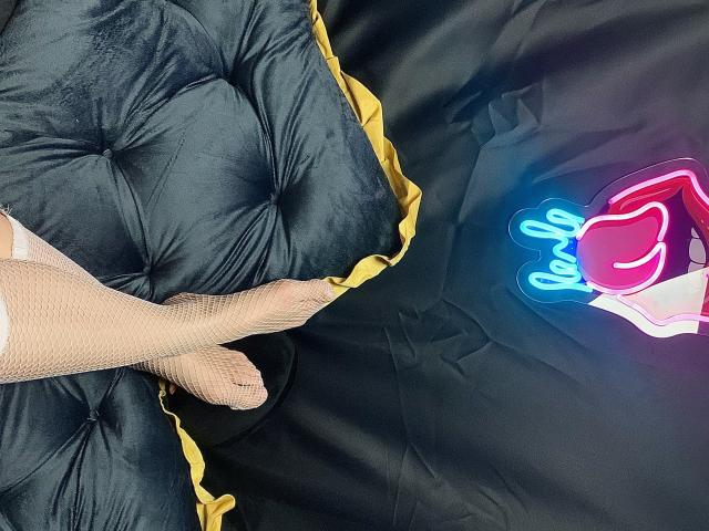 Le pied de DivineBlondee, portant des bas en filet, repose sur un lit noir en velours, avec des touffes et un bord jaune. Un panneau en forme de cœur, de couleur rose néon et bleu, se trouve à droite..