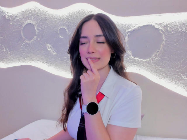 Maria, con capelli scuri, camicia bianca, cravatta rossa e orologio da polso nero, sorride, toccandosi il dito alle labbra, mostrando un'espressione carina e timida, con il seno visibile..