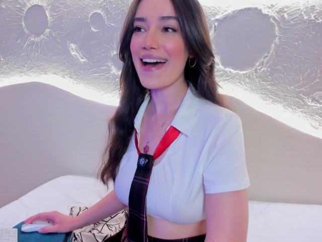 MariaLoft with long brown hair, smiling, wearing a white shirt with red collar and black tie, holding a blue object, her tits slightly visible, in a brightly lit room.
