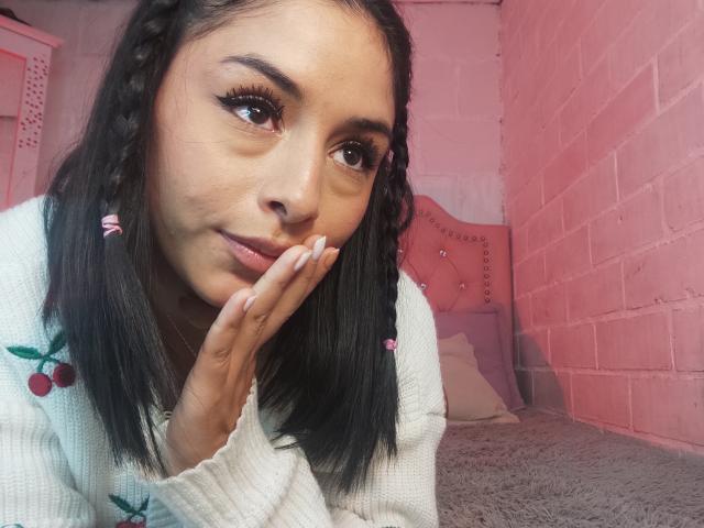 EmilyVv with black braided hair, pink earrings, and white sweater, resting her hand on her face, showing off long eyelashes, pink lips, and a cute smile.