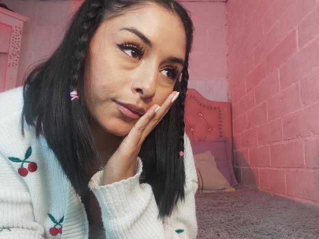 EmilyVv with long black braided hair, light brown skin, and pink nails, wearing a white knitted sweater with cherry patterns, resting her hand on her cheek, smiling.