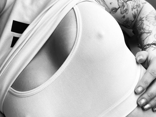 SujannaVarma's large, round boobs in a tight, white tank top, with a tattooed arm and glittery nails, shown in a black-and-white photo. Boob nipple slightly visible.