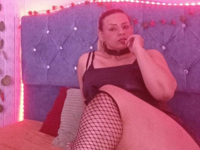 KathaleyaGrayyAss zit op een bed, gekleed in een zwarte jurk, visnetkousen en een choker, met haar vinger in haar mond, en toont haar vagina en borsten..