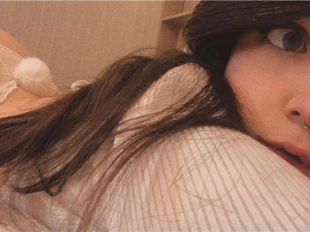 Das Gesicht von IrishReversed, mit großen Augen und langen braunen Haaren, trägt ein weißes Rippshirt und einen flauschigen weißen BH mit einem Pompon im Hintergrund..