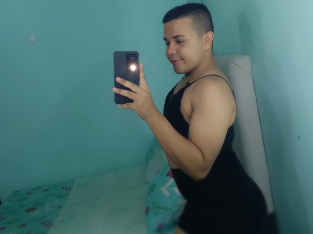 EmilyJedet, avec ses cheveux courts et foncés, ses bras musclés, un débardeur noir, prenant un selfie devant un miroir, souriante, tenant un téléphone avec un flash, debout dans une pièce bleu turquoise, un lit est visible..