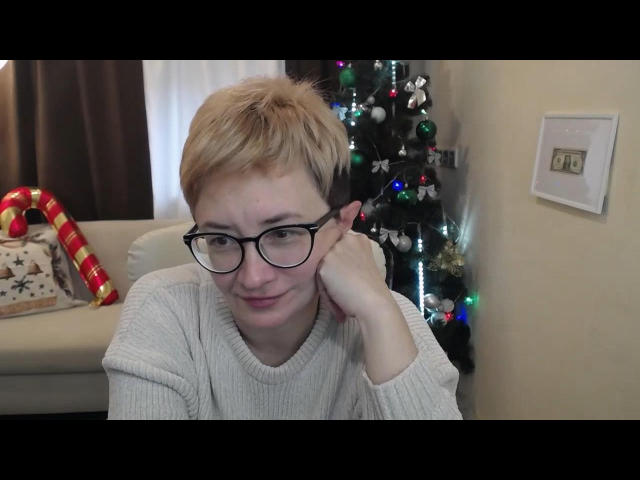 ChristinaRainforce with short blonde hair, black glasses, and a white sweater, resting her cheek on her hand, smiling subtly. Christmas tree with colorful lights in background.