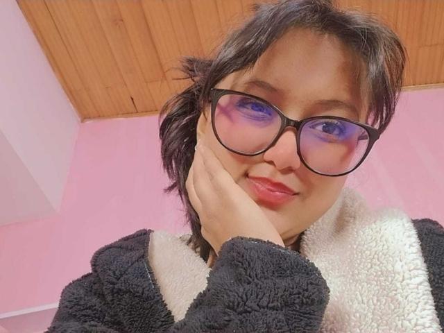 AzulMoon, con i capelli neri corti, gli occhiali, gli occhi di colore rosa e il rossetto rosso, che si tocca la guancia, indossa un cappotto morbido bianco e nero e sorride..