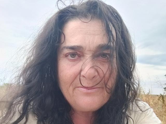 SaraElla's face with wavy black hair, a slight smirk, and a nose-poke by a fly. She has wrinkled skin, brown eyes, and is wearing a light-colored shirt.