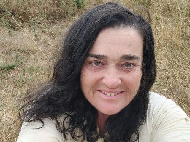 SaraElla with long, wavy black hair, smiling with slightly wrinkled face, green eyes, and a small nose. She's wearing a white shirt, standing in dry grass.