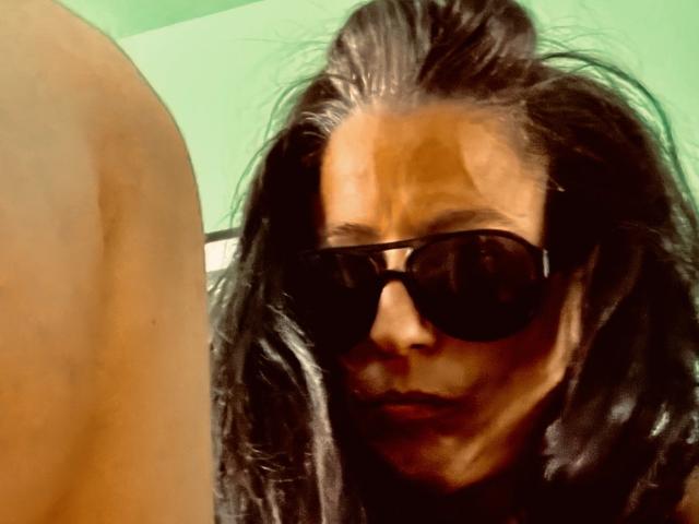 LorenaM with dark, wavy hair, wearing black sunglasses, looking to the side with a serious expression, upper torso and arm in the foreground.