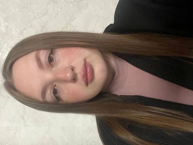 GirlPlayer-hot has long brown hair, pink turtleneck, black jacket, nose ring, freckles, and pink lips, looking at camera with subtle smile, light skin, and brown eyes.
