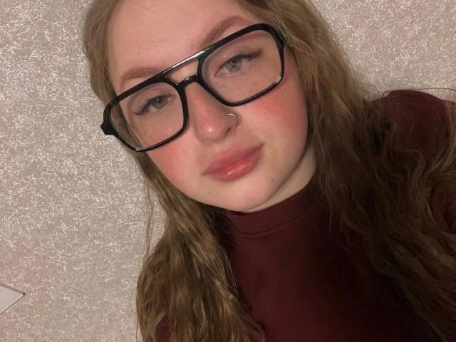 GirlPlayer-hot with long brown hair, wearing black glasses, a nose ring, and a burgundy turtleneck, has pinkish lips and a slightly flushed face.