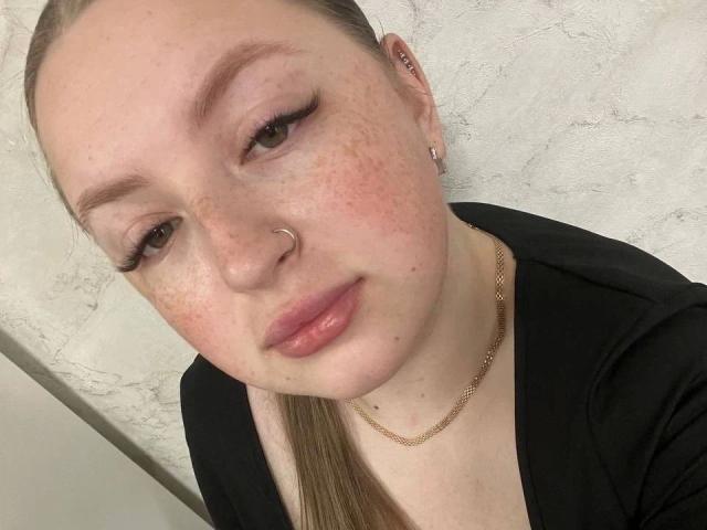 GirlPlayer-hot has a nose ring, freckled face, and brown eyes with long eyelashes. She's wearing a black top and a gold chain, looking slightly to the side.