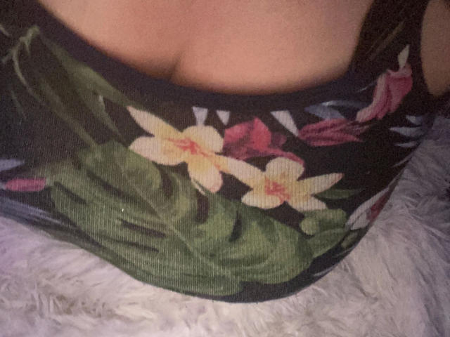Melli-hot's boobs, covered in a floral-patterned, black, ribbed tank top, with visible cleavage, sitting on a white, fluffy surface.