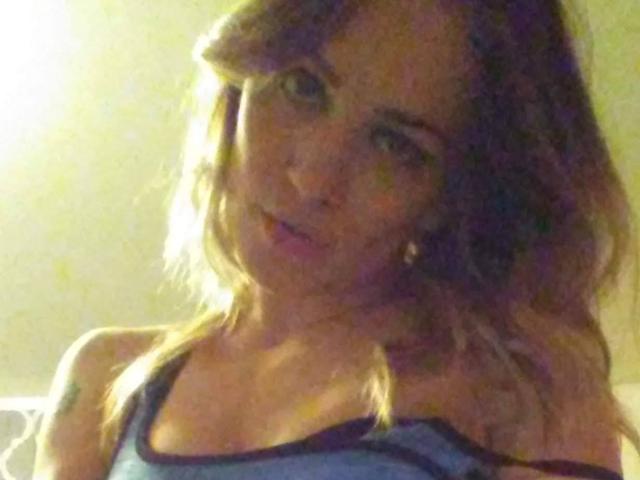 PhoenixPaulline with wavy brown hair, blue tank top, and subtle smile, looking slightly to the side, showing her small tits and a tattoo on her right shoulder.