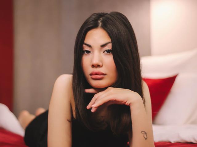MonicaLuxe mit langen braunen Haaren, vollen Lippen und einem Tattoo am rechten Handgelenk, liegt auf einem Bett, den Kopf in der Hand, und trägt ein schwarzes Kleid..