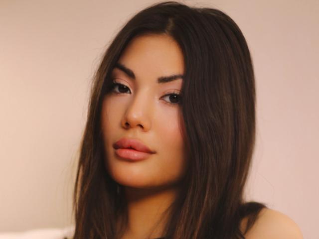 MonicaLuxe a de longs cheveux bruns, des lèvres pleines roses, et un joli visage avec des sourcils foncés et des yeux marron. Elle regarde légèrement sur le côté avec une expression neutre..