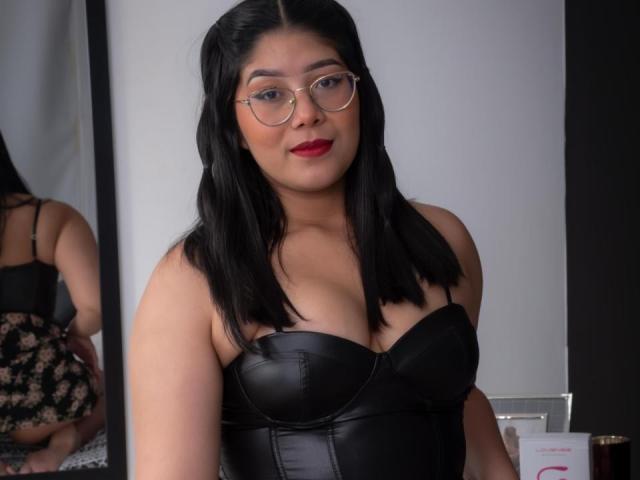 AlexaKiim with black leather bustier, showing her tits, wearing red lipstick and glasses, black wavy hair, mirror reflection of her ass, standing in front of a white wall.