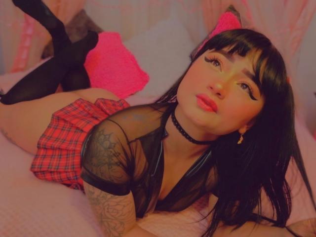 AnnieGrey with black hair, red plaid skirt, black mesh top, and black thigh-highs, lying on pink bed, showing tattoos on right arm, wearing choker, with pink pillow.